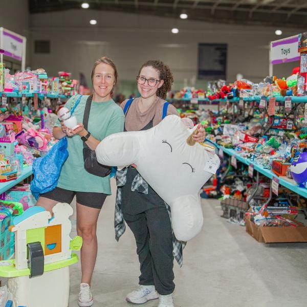 two smiling women holding toys and standing between tables of toys